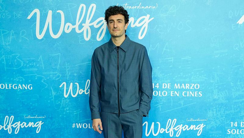 Actor con traje azul en photocall de estreno cinematográfico. Fondo azul claro con título de la película, fecha y hashtag.