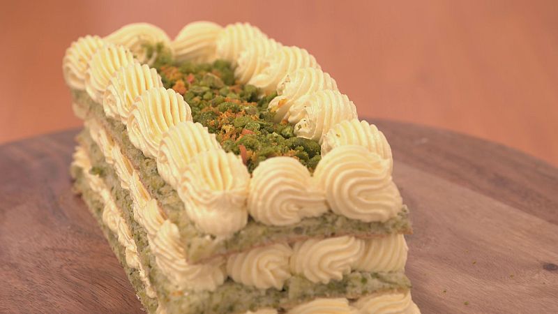 Bizcocho rectangular multicapa con crema, decorado con migas verdes y naranja confitada, sobre tabla redonda de madera.