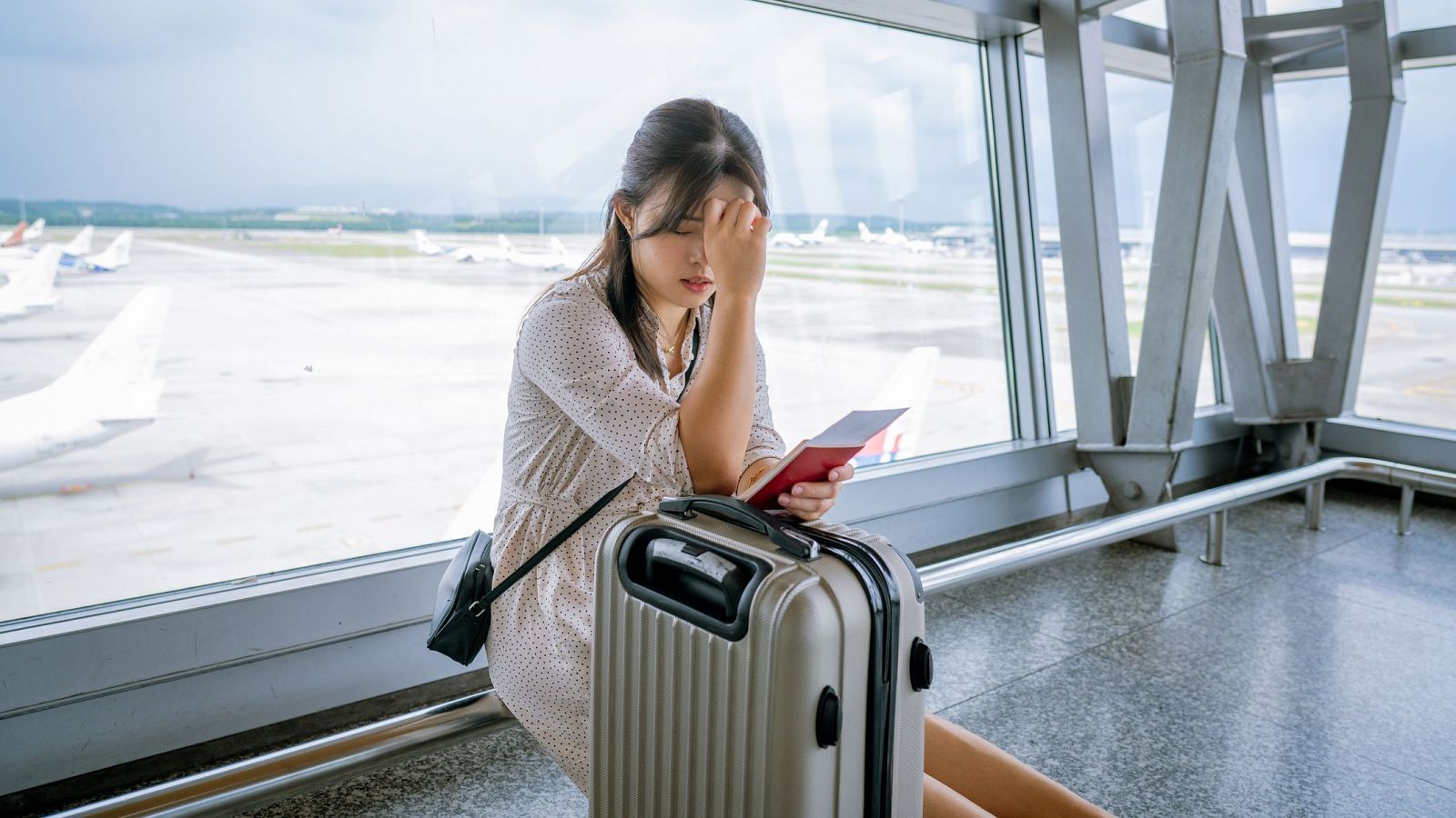 Mujer joven en sala de espera de aeropuerto, con expresión de ansiedad, sosteniendo pasaporte y billete.  Maletas pequeñas a su lado.