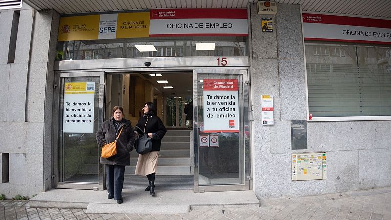 Entrada de oficina SEPE en Madrid (número 15).  Dos mujeres salen; una con abrigo marrón oscuro y bolso naranja, la otra con abrigo negro y falda beige. Rótulos indican 'Oficina de Empleo' y 'Oficina de Prestaciones'.