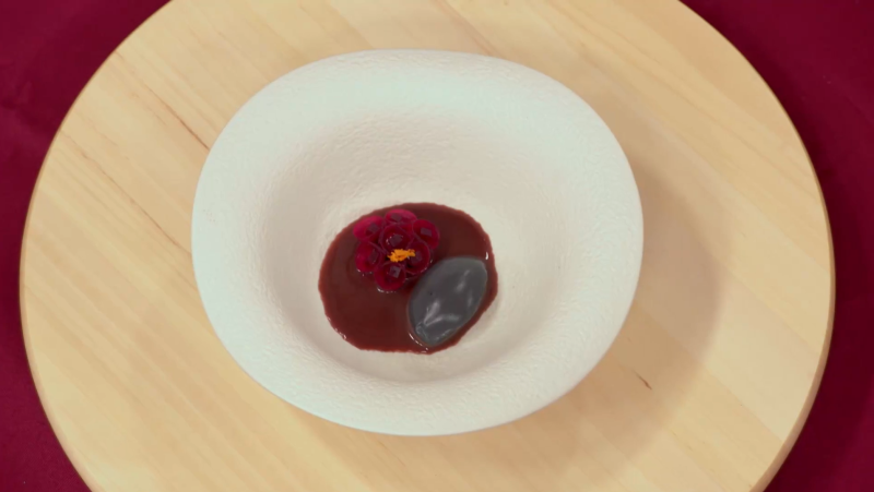 Postre de remolacha en forma de flor sobre salsa oscura y cremoso de ajo negro, presentado en plato blanco sobre base de madera.