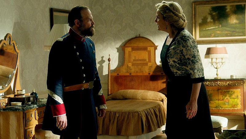 Hombre con uniforme militar y barba conversa con una mujer en un dormitorio con cama, tocador y cuadros.  Ambas expresiones sugieren una discusión seria.