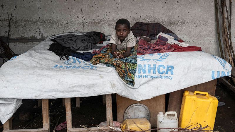 Niño refugiado sentado sobre una cama improvisada con lona del ACNUR en una habitación con daños. Lleva una camiseta clara y su expresión es seria.