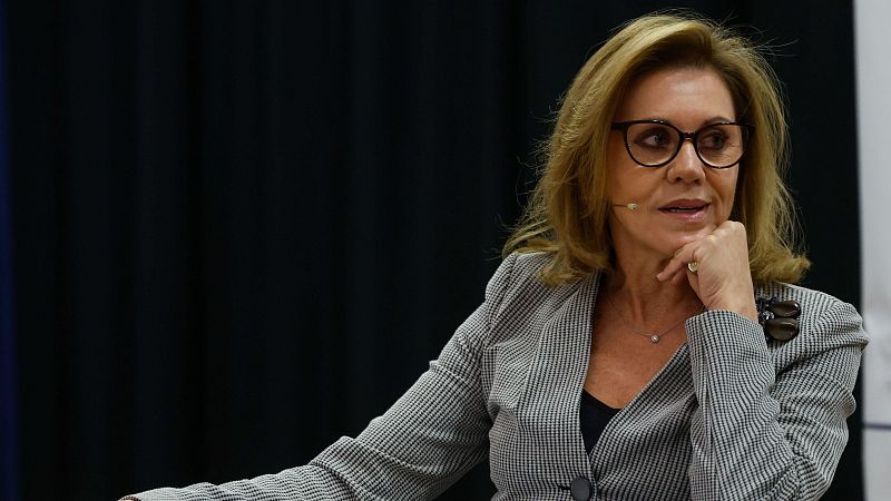 Imagen de María Dolores Cospedal con chaqueta de cuadros, gafas y expresión seria; parece una entrevista o evento público.