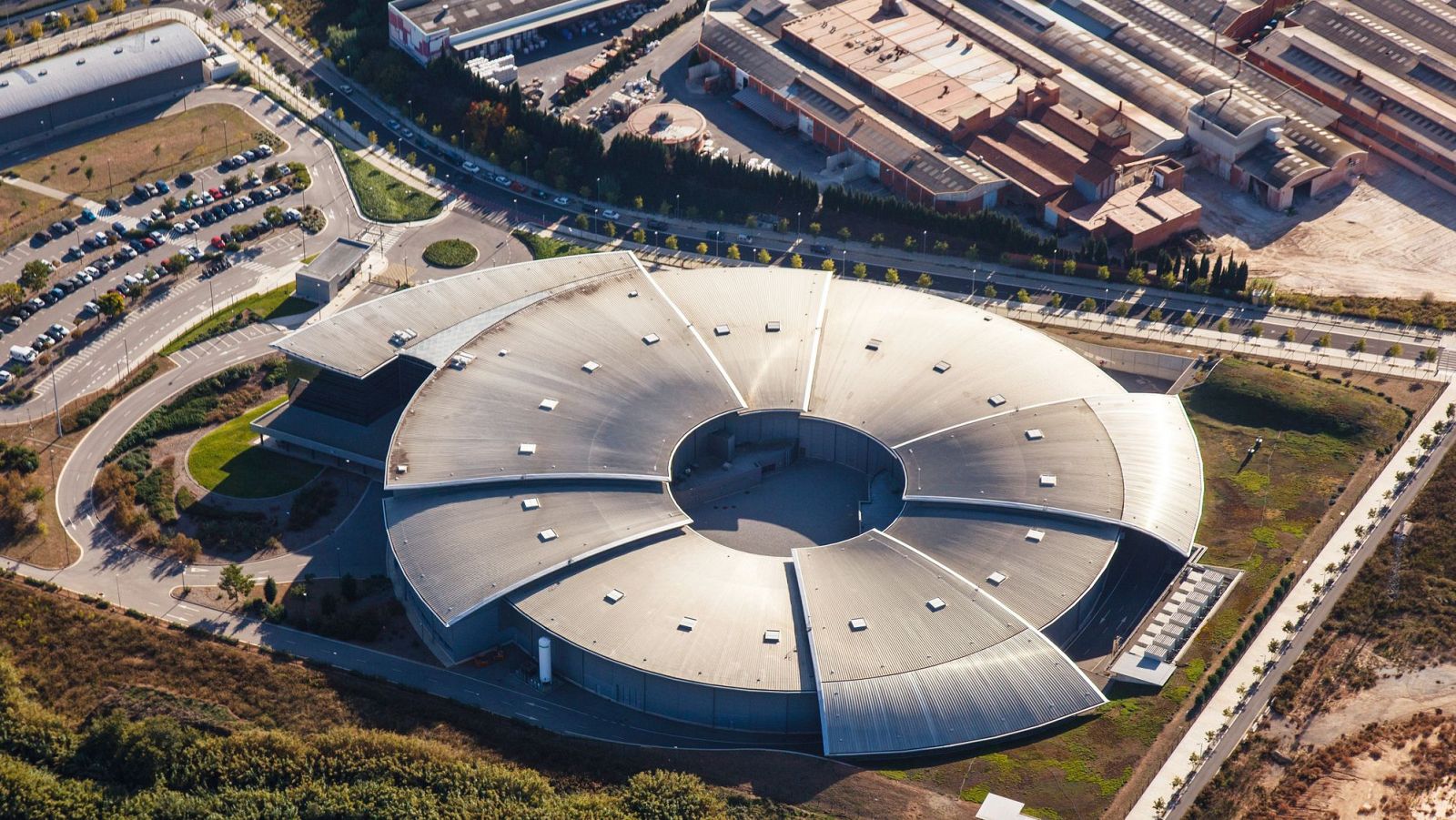 Vista aérea del Sincrotrón Alba: edificio circular con secciones petaloides, aparcamiento y construcciones adyacentes.