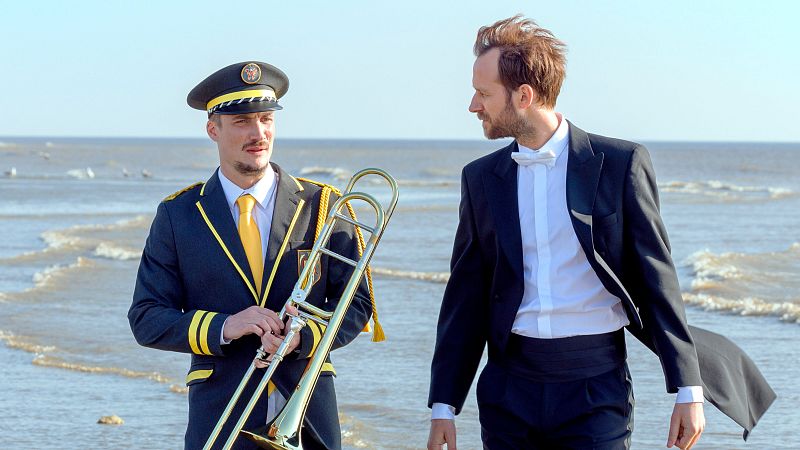 Dos hombres, uno en uniforme y trombón, otro en esmoquin, pasean por una playa soleada con mar en calma.