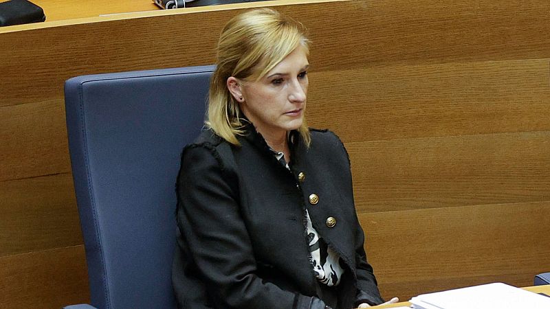 Consejera sentada en pleno, en sala con paneles de madera, viste chaqueta negra y blusa estampada, expresión seria, documentos en mesa.