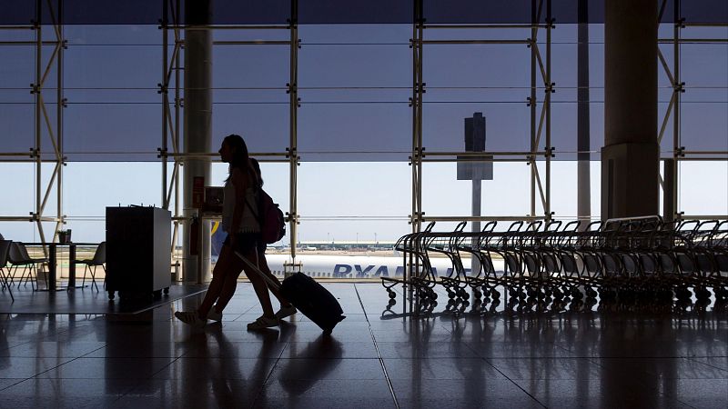 Siluetas de dos personas caminando en un aeropuerto, una con maleta; al fondo, un avión y carritos de equipaje se ven a través de ventanas.  Imagen en contraluz.