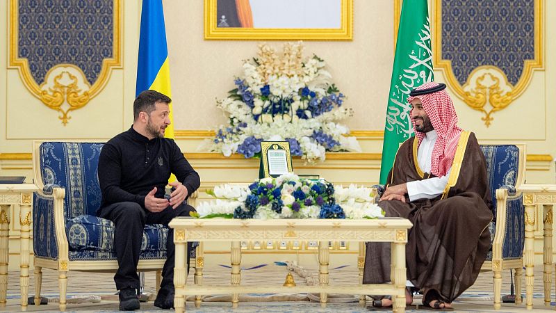 Zelenskyy y Bin Salman se reúnen en una sala palaciega. Él viste ropa oscura; él, túnica tradicional.  Sobre la mesa hay un documento y flores.