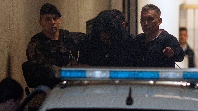 Detención de un hombre con la cabeza tapada, escoltado por un policía uniformado y otro civil. Vehículo policial con luces azules visibles.