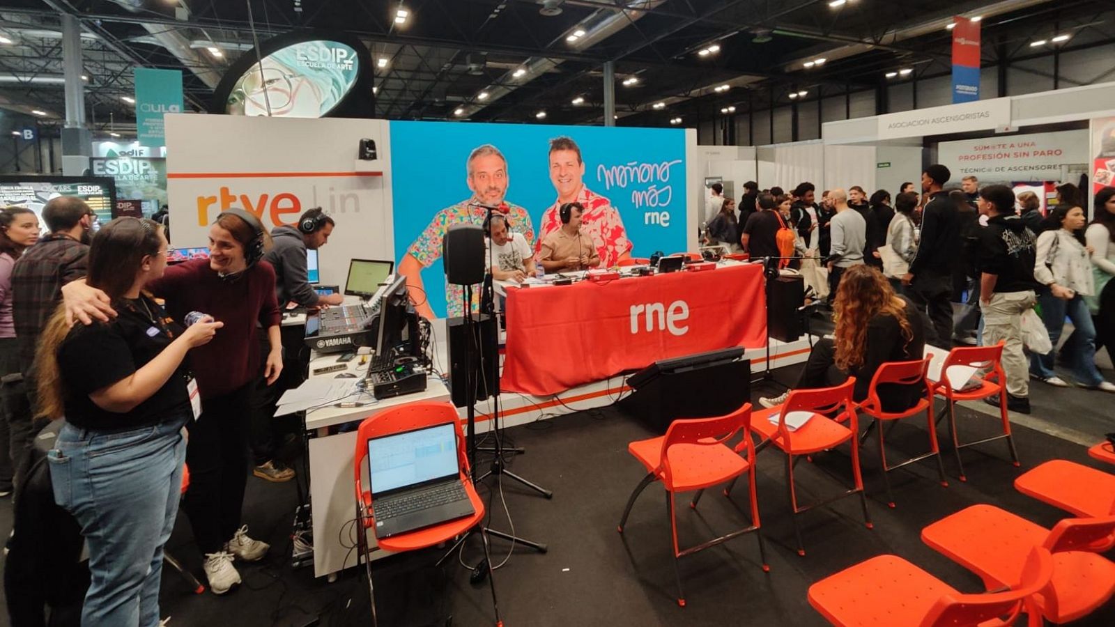 Stand de RTVE en un evento, con equipo de sonido, mesa con logo RNE, panel con logo RTVE y fotos de presentadores.  Técnico trabajando, dos mujeres conversando.