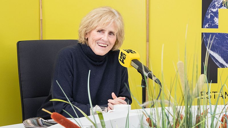 Mercedes Milá en entrevista para el podcast "HOPE!", sonriendo y vestida con un jersey azul oscuro.  Micrófono con logo visible.