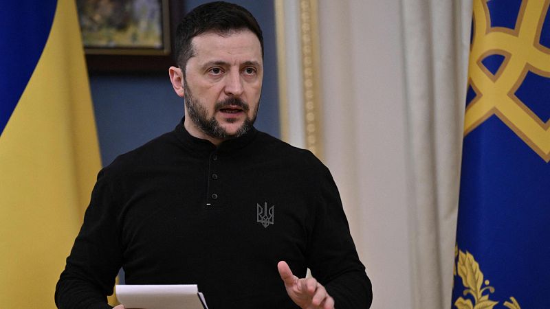 Comparecencia del presidente de Ucrania, vestido con jersey negro y escudo nacional bordado, hablando y gesticulando con papeles en la mano, ante dos banderas ucranianas.