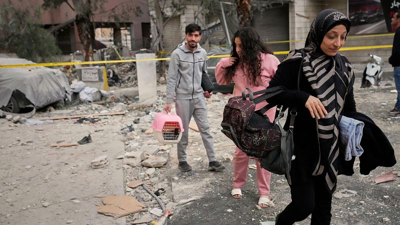 Escombros en una calle tras un bombardeo. Tres personas, una con jaula rosa y otra con hiyab, se alejan de la zona acordonada con cinta amarilla.
