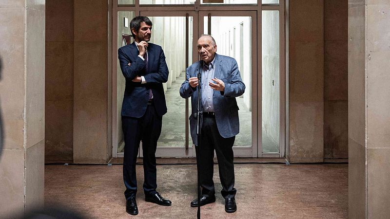 El ministro de Cultura y el presidente de la Academia de Cine conversan; uno habla por micrófono, el otro escucha con los brazos cruzados. Ambos visten formalmente.