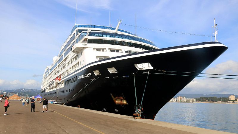 Proa de un crucero atracado, con varios pisos y ventanas visibles. Personas caminan por el muelle cercano.  Se observa parte de la ciudad al fondo.