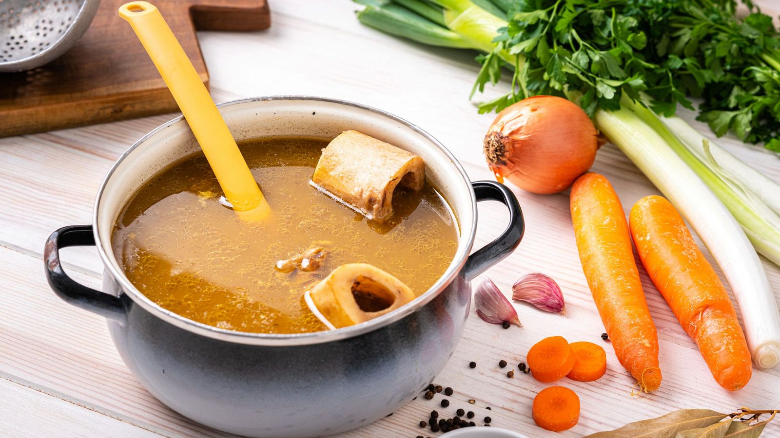 Caldo de huesos en olla gris oscura con asas negras y cuchara amarilla; ingredientes frescos alrededor: zanahorias, cebolla, puerro, perejil, ajo, pimienta y laurel.