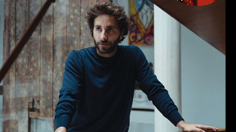 Hombre con barba y jersey azul, apoyado en barandilla, fondo con pared de madera y arte abstracto.  Imagen promocional relacionada con "Las noches del Monumental".