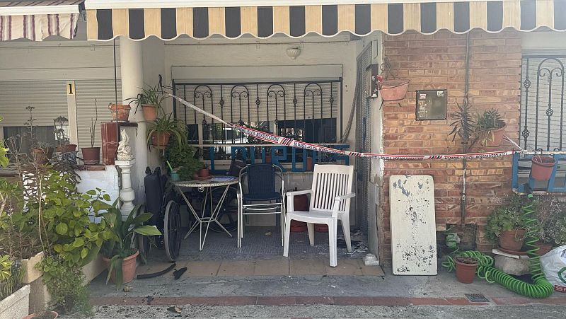 Porche acordonado con cinta policial, mostrando daños en la puerta y ventana.  Muebles de plástico, plantas y una manguera presentes en la escena.