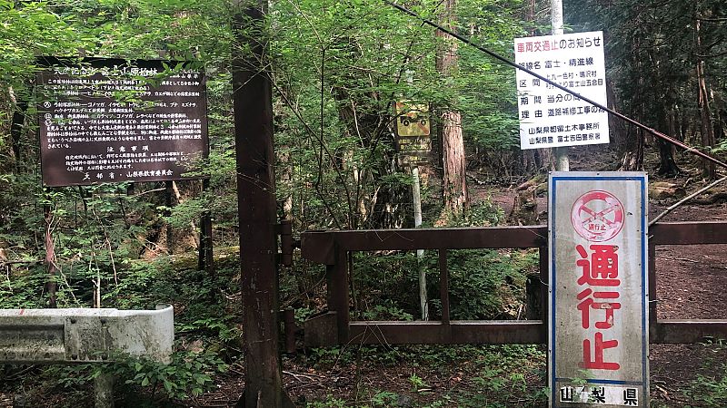 Entrada a zona boscosa restringida con barrera metálica y señales indicando cierre de carretera y prohibición de paso.  Se observa texto en japonés en una señal y símbolos de prohibición.