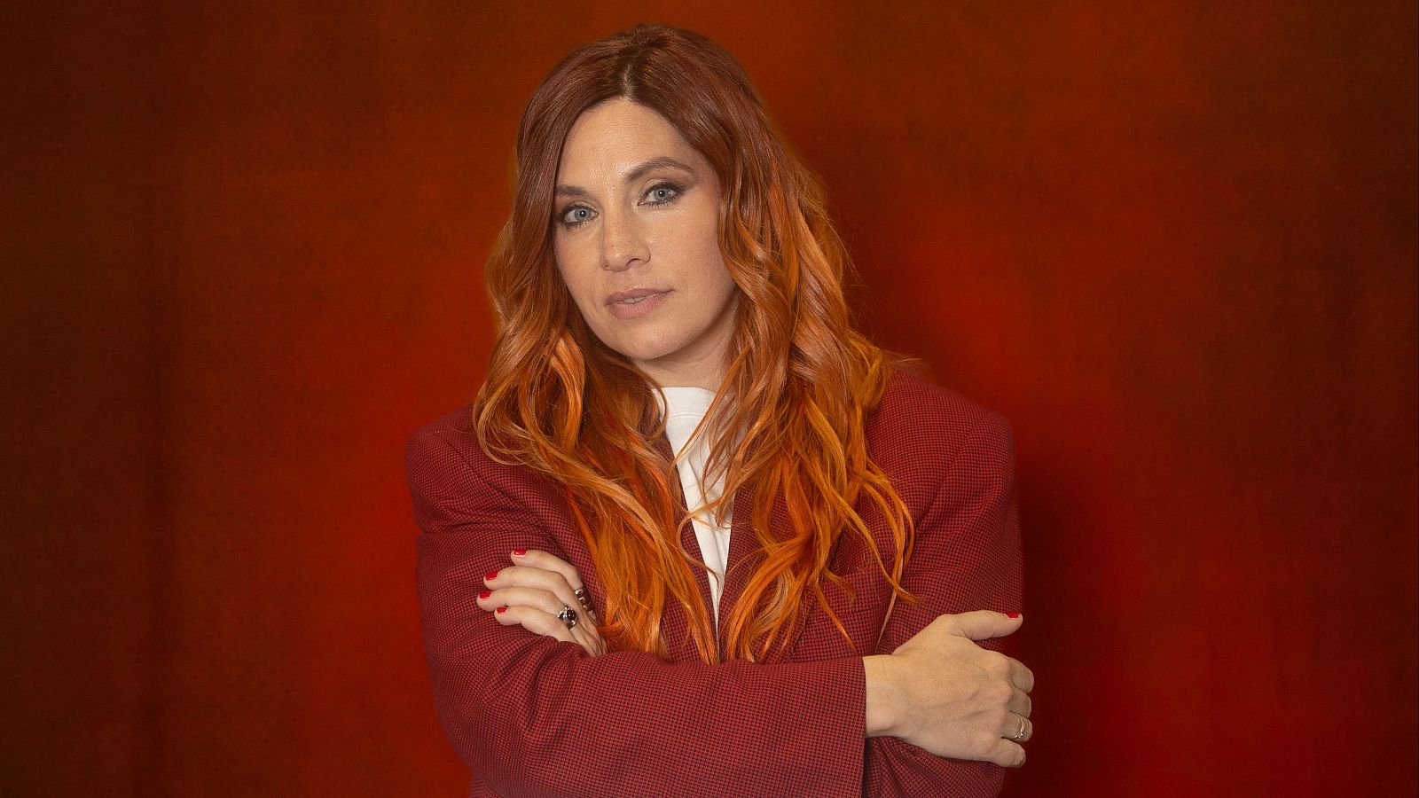 Leire Martínez posa con americana granate y pelo rojizo sobre fondo rojo oscuro. Brazos cruzados, uñas pintadas de rojo oscuro.