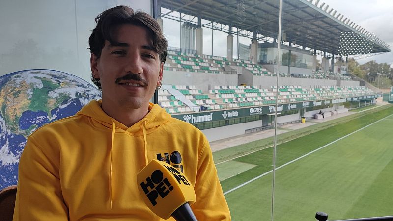 Entrevista a Héctor Bellerín en el podcast "Hope!".  El futbolista viste sudadera amarilla con el logo y usa un micrófono amarillo con el mismo logo. Fondo con vista a un campo de fútbol.
