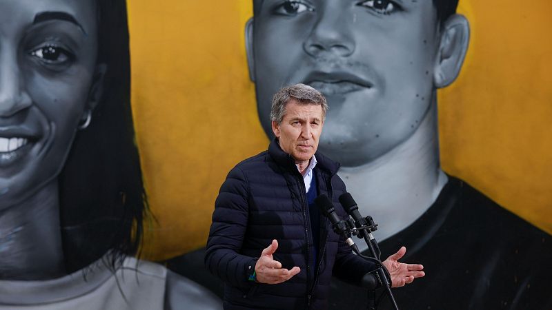 El presidente del PP, con chaqueta azul, ofrece declaraciones a la prensa, gesticulando ante dos micrófonos. Detrás, un mural con dos rostros en blanco y negro.