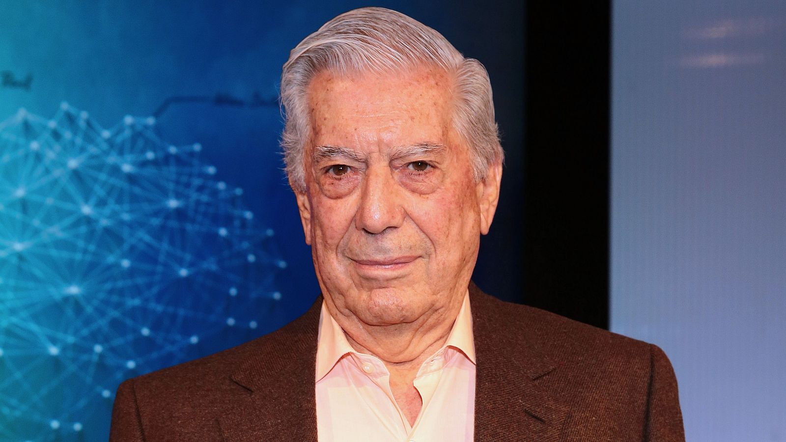 Mario Vargas Llosa en foto de archivo (2018).  Expresíon seria, pelo canoso hacia atrás, camisa clara y chaqueta marrón. Fondo abstracto.