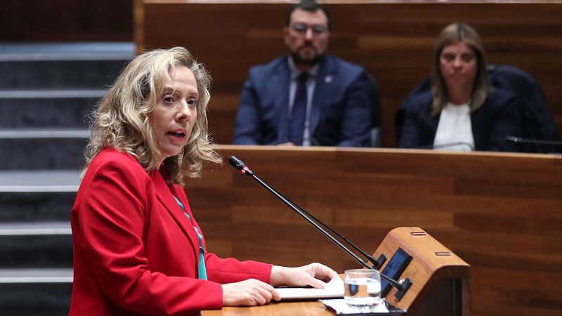 Mujer en chaqueta roja habla en atril de madera ante dos personas sentadas en sala con paneles.  Micrófono y vaso de agua presentes.