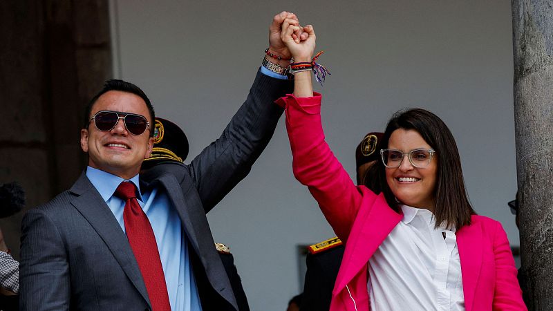 Daniel Noboa y María José Pinto celebran su victoria electoral; él en traje gris, ella con chaqueta rosa, ambos con gafas de sol y sonrisas.  Uniformes militares al fondo.