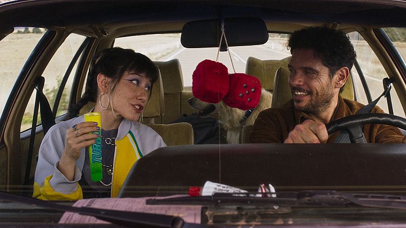 Dos personas en un coche, una mujer con chaqueta deportiva y un hombre conduciendo.  Un perro en el asiento trasero. Objetos en el salpicadero.