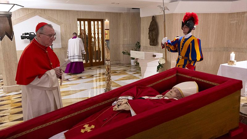 Constatación de la muerte del Papa en la Capilla de Santa Marta.  Un cardenal y un guardia suizo rodean el féretro rojo.  Se observa una cámara de televisión.