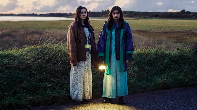 Dos chicas, una con linterna, caminan juntas por un camino al atardecer, junto a un paisaje con agua y árboles.