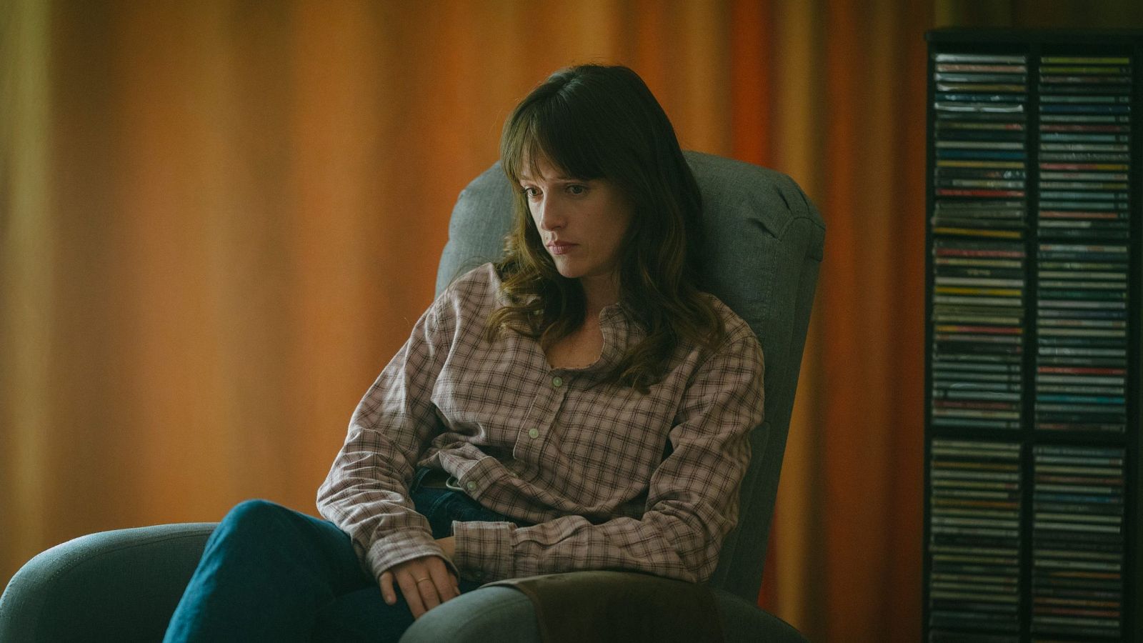 Actriz sentada en sillón gris, camisa a cuadros, vaqueros oscuros, expresión seria. Ambiente íntimo y melancólico.