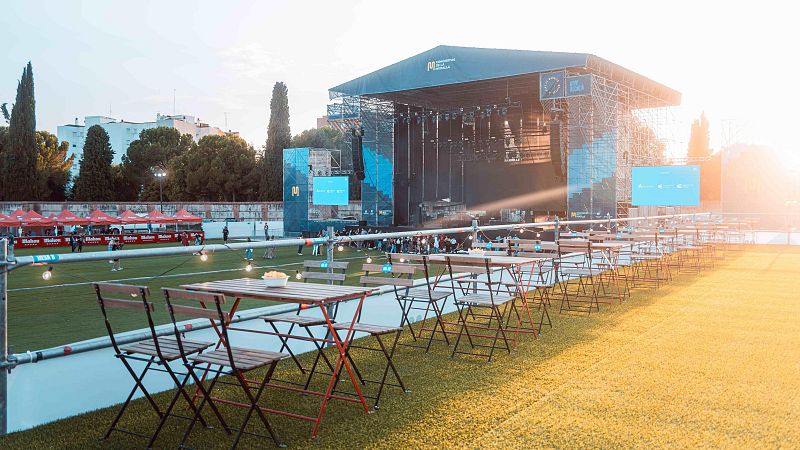 Mesas y sillas plegables en zona de descanso festivalera, separadas por valla metálica con luces de un escenario con logo visible. Atardecer.
