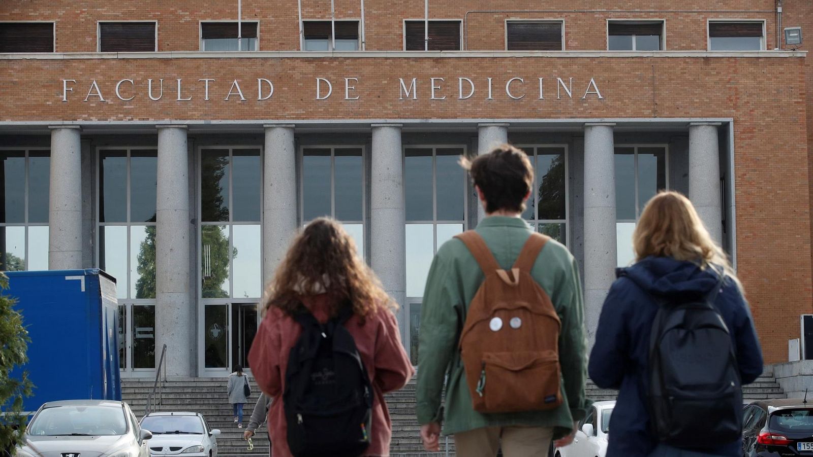 Fachada de facultad con tres estudiantes con mochilas caminando hacia la entrada.  Se aprecian detalles de sus chaquetas (rojiza, verde claro, azul marino) y un camión de mudanzas en el fondo.
