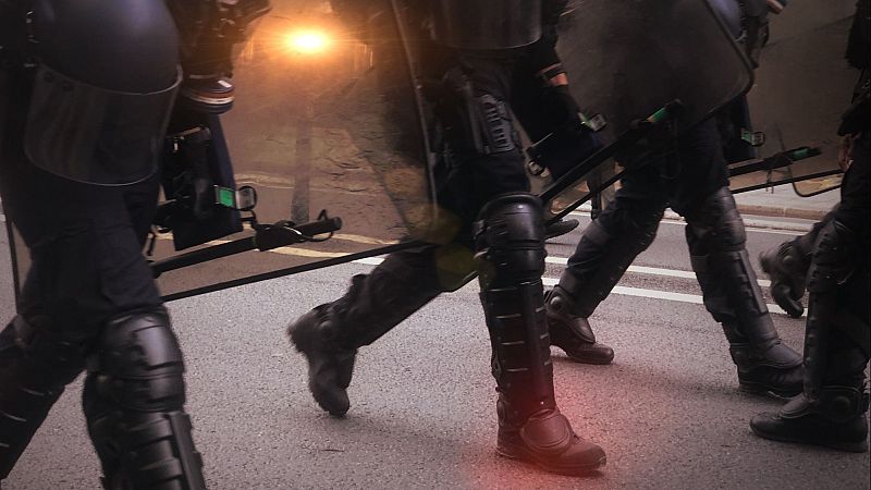Un grupo de policías antidisturbios, con equipo completo (cascos, protectores y escudos), caminan a paso ligero.  Uniformes oscuros, fondo brillante.