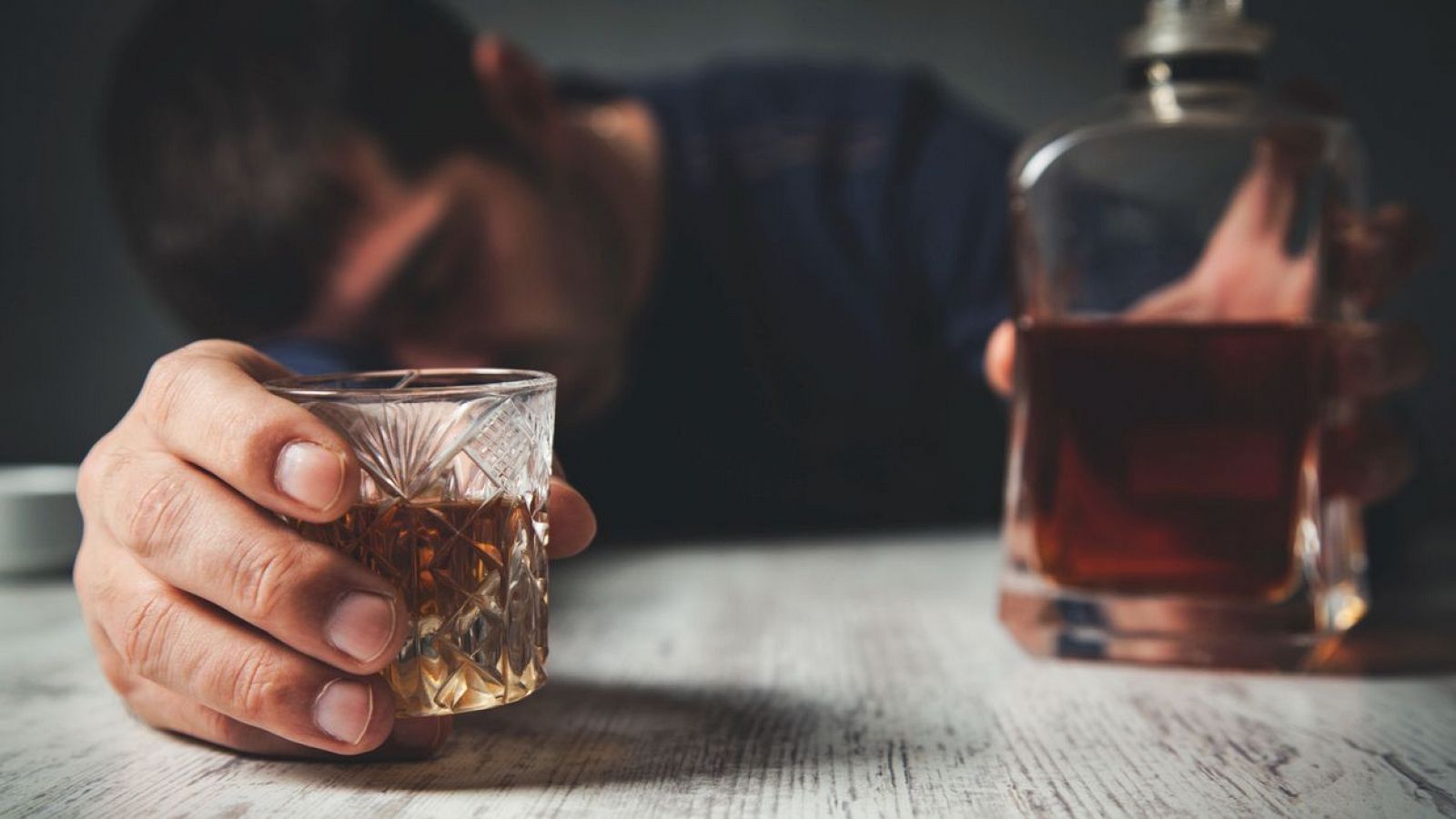 Hombre ebrio con vaso de bebida alcohólica en mano, apoyado en una mesa con una botella de licor visible. Imagen que refleja los efectos del alcoholismo.