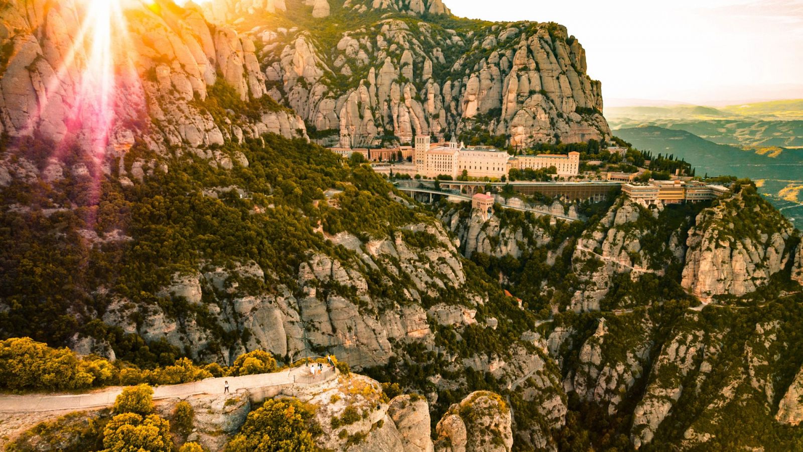 Vista aérea de Montserrat con sus rocas irregulares, vegetación y el monasterio.  Personas caminan por un sendero montañoso. Cielo con tonos naranja y rosa.
