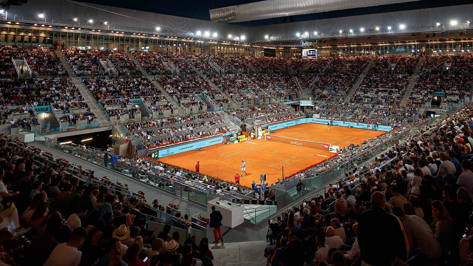 Partido de tenis nocturno en pista de tierra batida, con jugadores, público y jueces. Iluminación artificial en un estadio lleno.