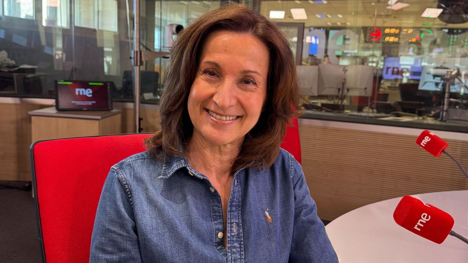 Mujer en estudio de radio con dos micrófonos, sonriendo a cámara.  Ambiente profesional con paredes de cristal.