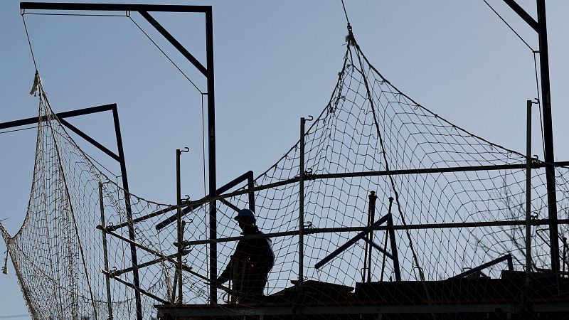 Trabajador en andamio con red de seguridad, manipulando herramientas. Cielo despejado al fondo.