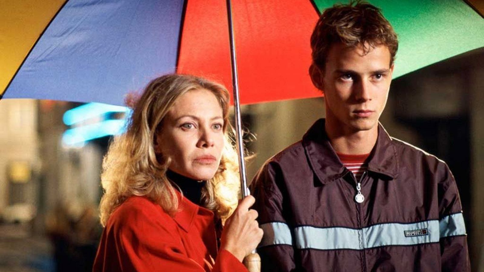 Escena nocturna de "Todo sobre mi madre" con Cecilia Roth (abrigo rojo) y Eloy Azorín (chaqueta marrón) bajo un paraguas multicolor.