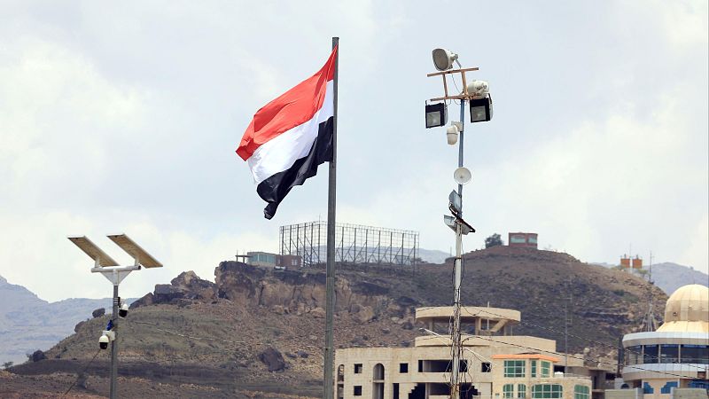 Bandera de Yemen ondeando en un mástil, con edificios y terreno montañoso al fondo. Varios postes con focos, antenas y paneles solares visibles.
