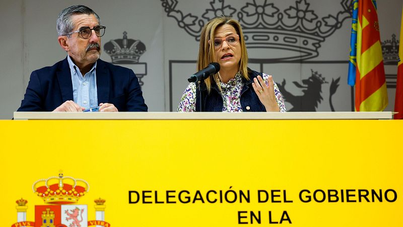 Declaraciones de Pilar Bernabé con micrófono en sala oficial con atril y banderas de España y la Comunidad Valenciana.