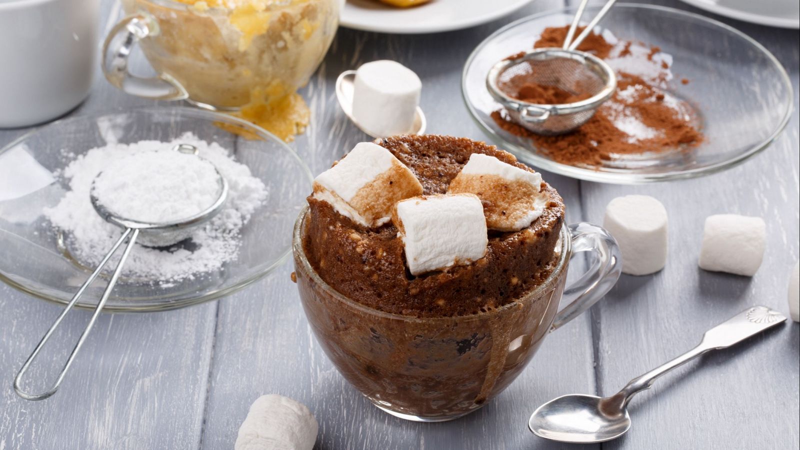 Bizcocho de chocolate húmedo con malvaviscos en taza, acompañado de ingredientes y utensilios de repostería sobre una mesa de madera.