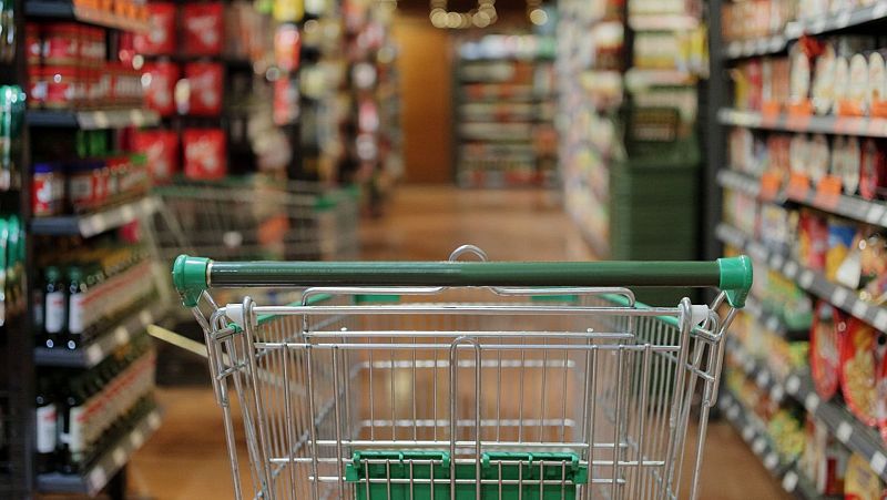 Carrito vacío en pasillo de supermercado con estantes repletos.  Ilustra noticia sobre incremento del 19% en gasto mensual de alimentación desde 2019.