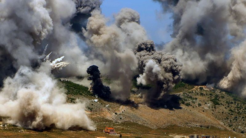 Paisaje montañoso con varias columnas de humo, un camino y pequeñas estructuras.  Nube de polvo indica explosión reciente. Perspectiva aérea.