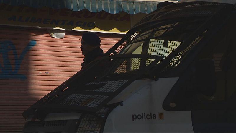 Agente policial junto a furgoneta con rejas; visible la palabra "policia" y bandera española. Fondo con persiana metálica.