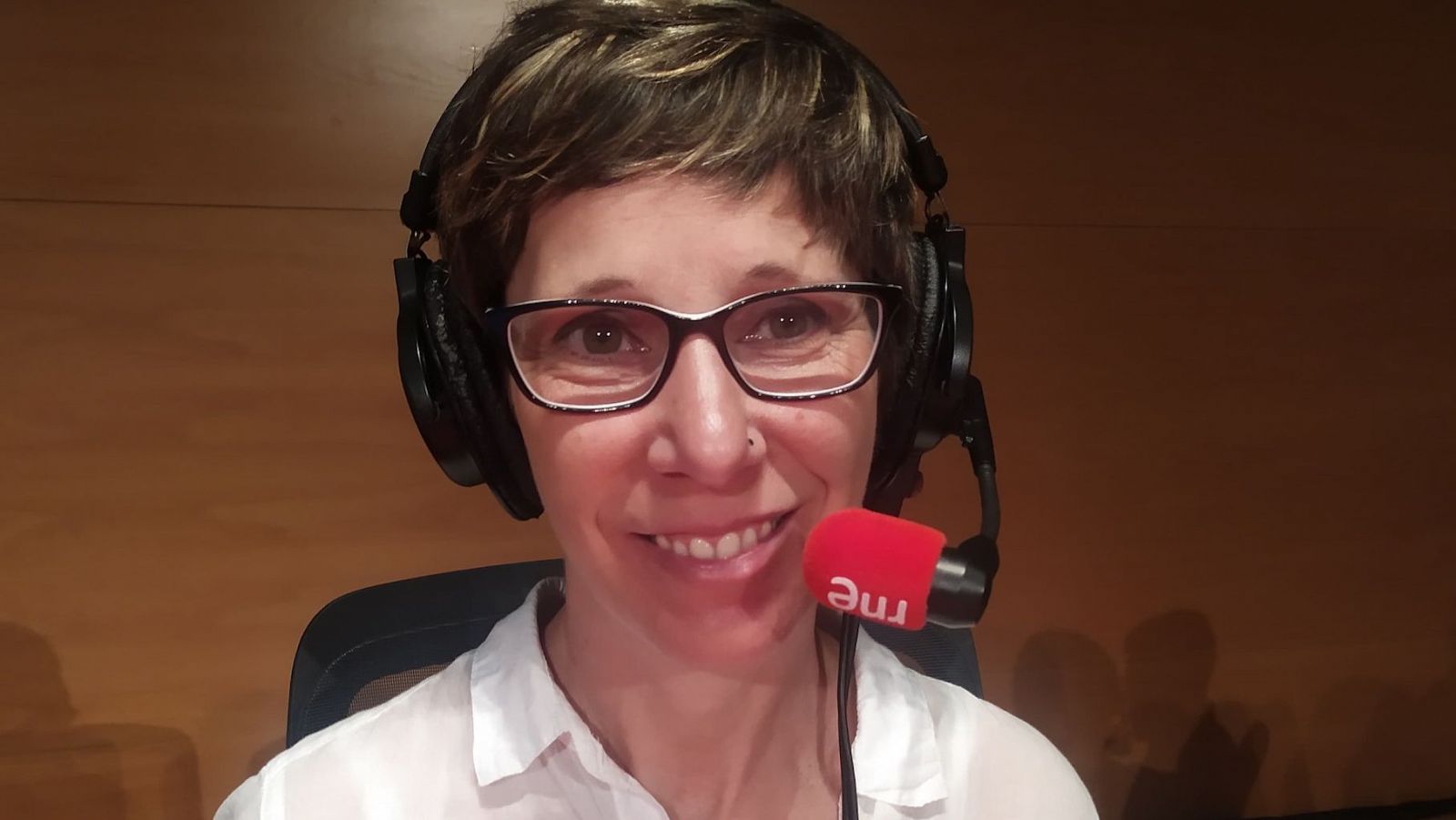 Locutora o periodista de RNE en estudio de radio, con auriculares y micrófono con protector rojo. Sonríe, viste camisa blanca y el fondo es marrón.